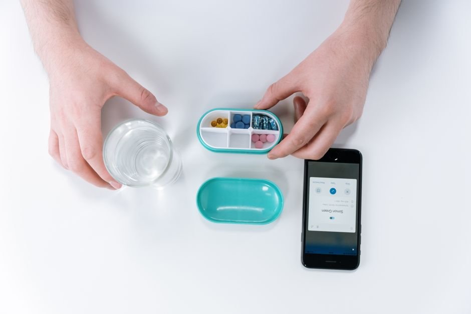 A person organizing pills in a compartmentalized container next to a smartphone and a glass of water, illustrating medication scheduling.